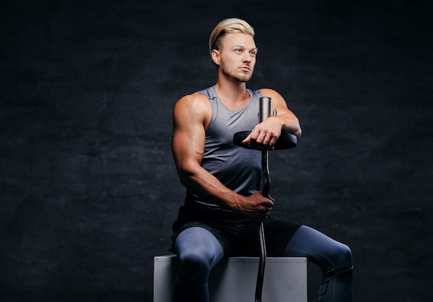 Un homme athlétique blond en tenue de sport est assis sur une boîte blanche et tient une barre.