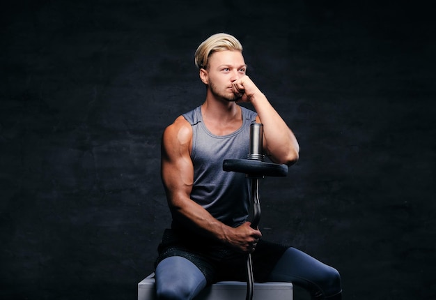 Un homme athlétique blond en tenue de sport est assis sur une boîte blanche et tient une barre.