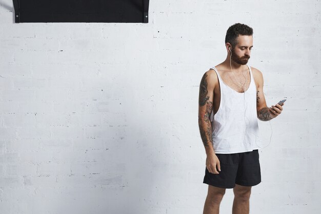 Homme athlétique brutal tatoué en t-shirt réservoir blanc regarde dans son smartphone, portant des écouteurs avant l'entraînement sur fond de brique blanche