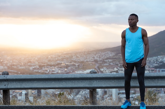 Un homme athlétique à la peau sombre porte des vêtements confortables et décontractés, se repose après un entraînement physique actif, aime les exercices physiques, se concentre sur la distance, a la peau foncée, les lèvres charnues. Copiez l'espace sur la vue de la nature