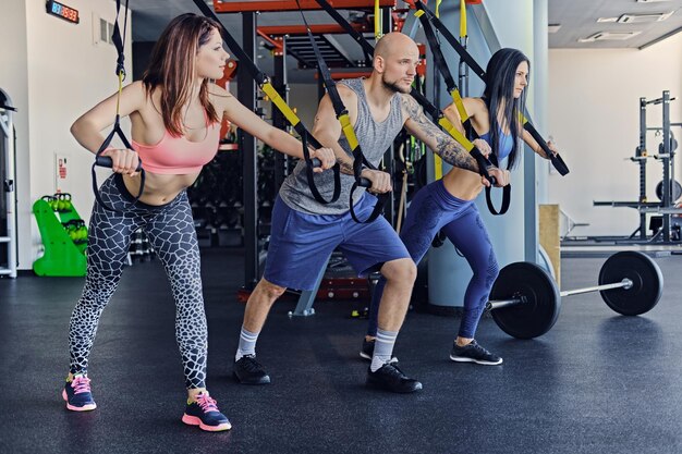 Homme athlétique tatoué et deux femmes sportives faisant des exercices de sangles trx.