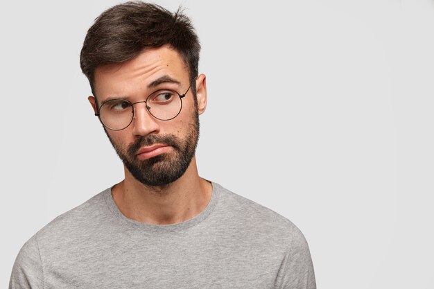 Un homme attrayant mal rasé pensif regarde pensivement de côté, a des pensées profondes à l'esprit, a des lunettes rondes, pose contre un mur blanc