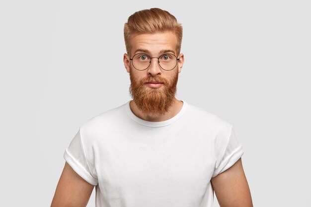 Homme au gingembre confiant avec une coiffure à la mode, porte des lunettes, regarde directement