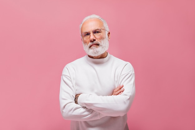 Homme aux cheveux blancs en pull et lunettes posant sur un mur rose