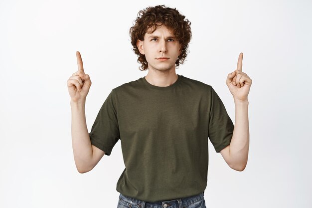 Homme aux cheveux bouclés agacé pointant les doigts vers le haut en regardant la publicité de lecture de logo avec l'expression du visage irrité sur fond blanc