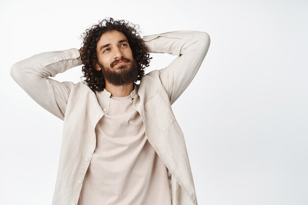 Homme aux cheveux bouclés insouciant tenant les mains derrière le dos relaxant et souriant allongé et reposant debout sur fond blanc