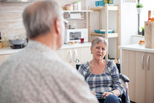 Homme aux cheveux gris discutant avec sa femme paralysée. Femme invalide à la retraite en fauteuil roulant ayant une conversation avec un vieux mari âgé dans la cuisine. Vieil homme parlant avec sa femme. Vivre avec une personne handicapée avec