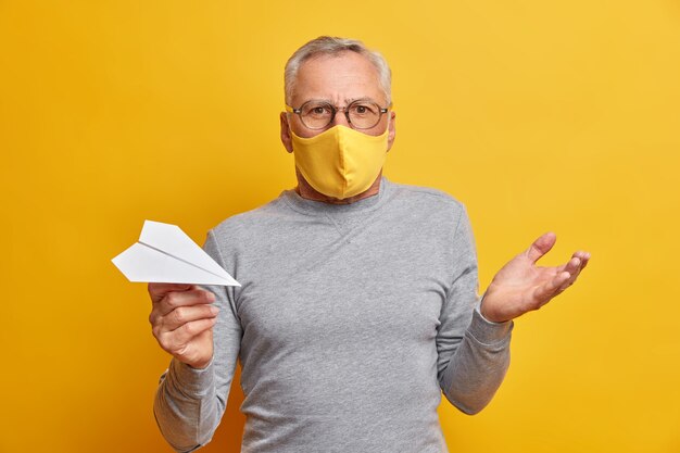 Un homme aux cheveux gris hésitant et perplexe lève la paume et se sent confus porte un masque jetable pour se protéger du coronavirus tient un avion en papier fait à la main isolé sur un mur jaune