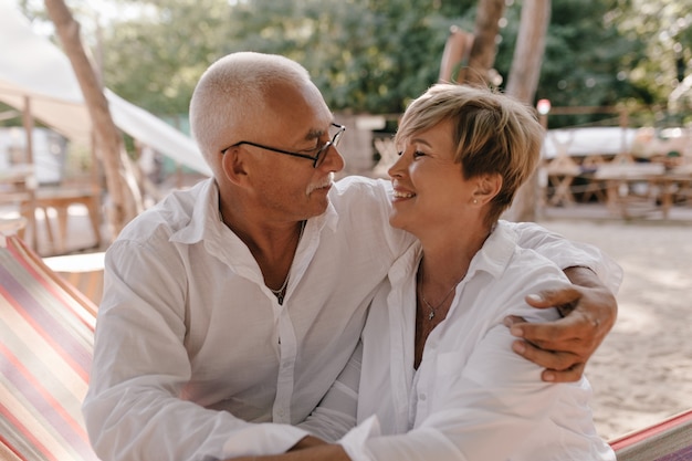 Homme aux cheveux gris avec des lunettes en chemise à manches longues étreignant et regardant femme souriante aux cheveux courts en chemisier blanc sur la plage.