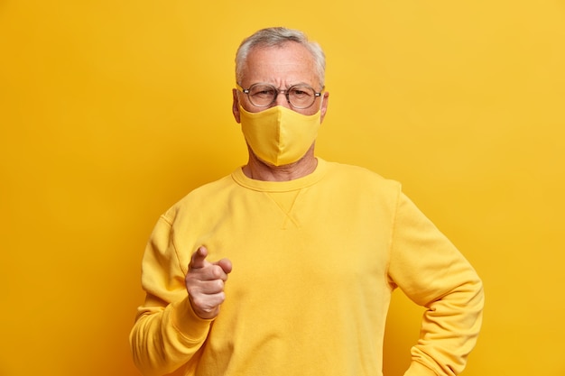 Photo gratuite homme aux cheveux gris sérieux regarde avec une expression stricte à l'avant des points d'index vers l'avant masque facial jaune weas comme protection contre le virus se trouve à l'intérieur