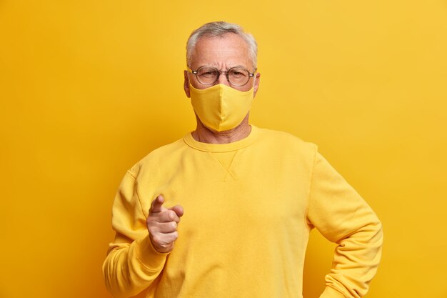 Homme aux cheveux gris sérieux regarde avec une expression stricte à l'avant des points d'index vers l'avant masque facial jaune weas comme protection contre le virus se trouve à l'intérieur