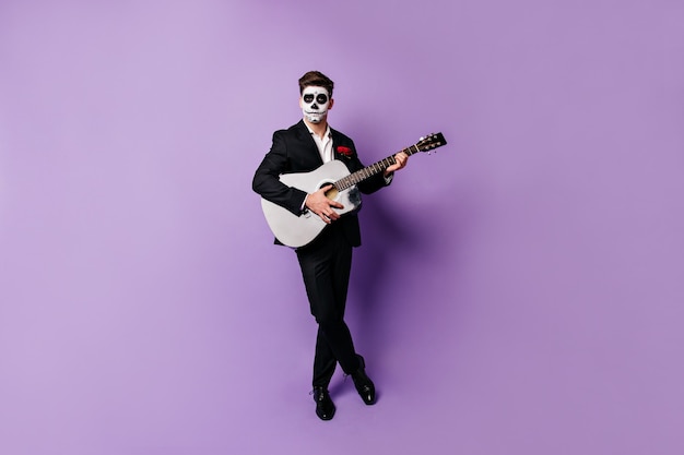 Homme aux cheveux noirs en costume élégant et visage peint en forme de crâne joue de la guitare, regardant dans la caméra avec un regard impassible.