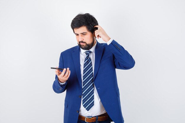 Homme aux cheveux noirs regardant le téléphone tout en se grattant la tête en chemise blanche, veste bleu foncé, cravate, vue de face.