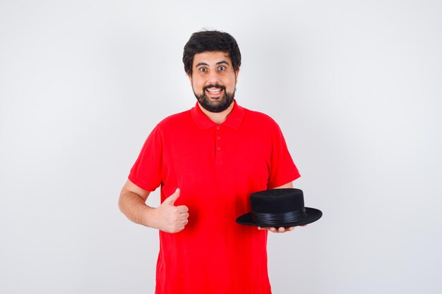 Homme aux cheveux noirs en t-shirt rouge tenant un chapeau tout en montrant le pouce vers le haut et l'air heureux, vue de face.