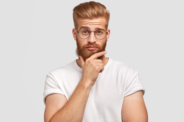 Homme aux cheveux rouges hésitant perplexe tient le menton et lève les sourcils avec une expression désemparée