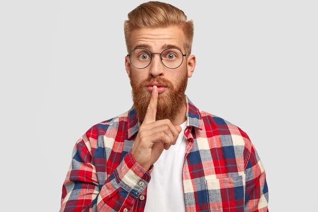 homme aux cheveux roux sérieux, a une barbe épaisse de gingembre, garde l'index sur les lèvres, regarde secrètement, a une expression stupéfaite