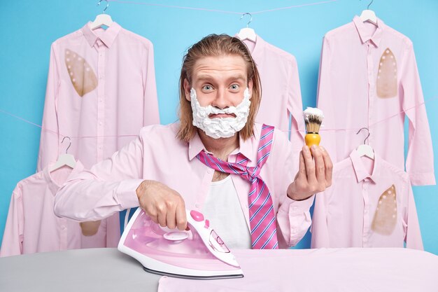 un homme aux cheveux roux utilise une brosse pour appliquer le gel à raser se tient près d'une planche à repasser des vêtements froissés des robes pour le travail