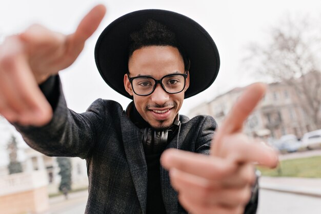 Homme aux yeux bruns de bonne humeur s'amusant pendant une séance photo en plein air. Photo d'un mec africain souriant dansant dans la rue par temps froid.