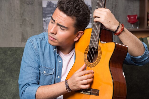 Homme aux yeux fermés tenant une belle guitare et assis sur un canapé. Photo de haute qualité