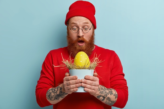 Homme avec barbe au gingembre portant des vêtements colorés et tenant l'oeuf de Pâques