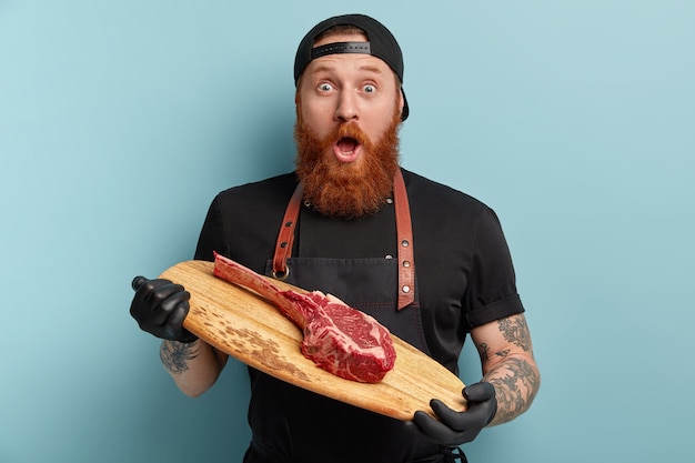 Homme avec barbe au gingembre en tablier et gants tenant une planche de bois avec un morceau de viande