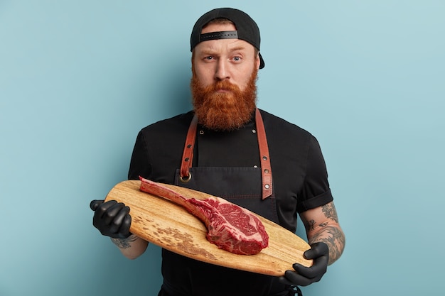 Homme avec barbe au gingembre en tablier et gants tenant une planche de bois avec un morceau de viande