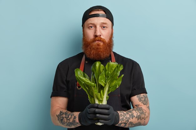 Homme avec barbe au gingembre en tablier et gants tenant une salade