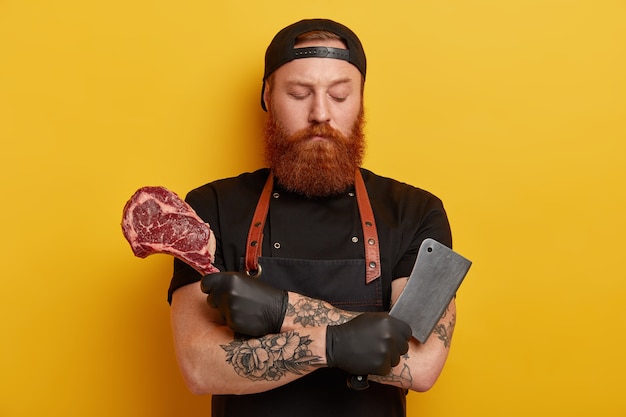 Homme avec barbe au gingembre en tablier et gants tenant la viande et le couteau