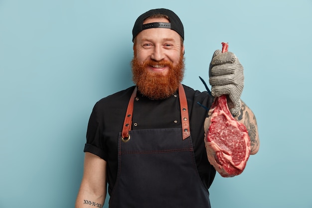 Homme avec barbe au gingembre en tablier et gants tenant de la viande