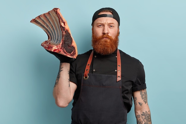 Homme avec barbe au gingembre en tablier et gants tenant de la viande