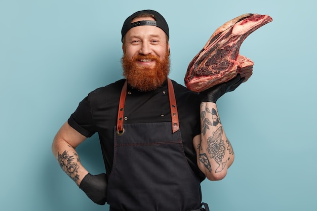 Homme avec barbe au gingembre en tablier et gants tenant de la viande