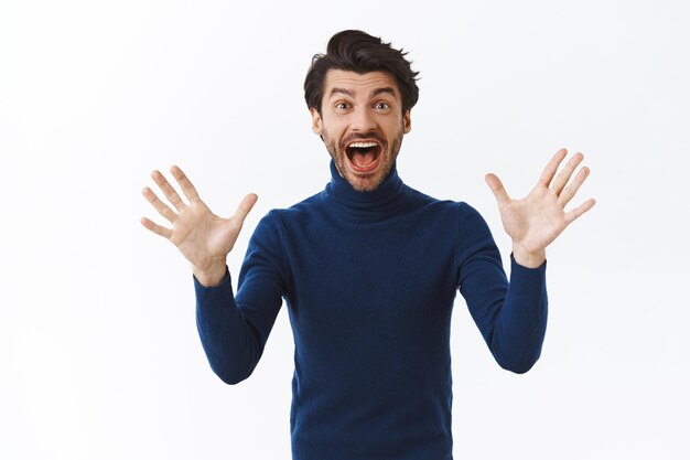 homme à la barbe, cheveux noirs, porte un pull à col haut, serrant les mains levées comme triomphant, célébrant d'excellentes nouvelles