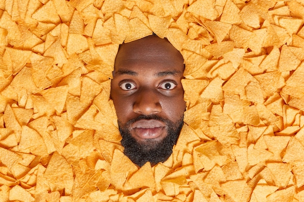 Photo gratuite un homme avec une barbe épaisse regarde impressionné entouré de chips croustillantes mange une collation malsaine consomme beaucoup de calories