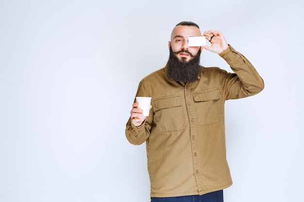 Homme à la barbe montrant sa carte de visite tout en tenant une tasse de café.