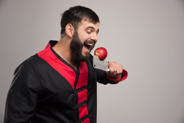 Homme à barbe mordant une pomme rouge sur un couteau.