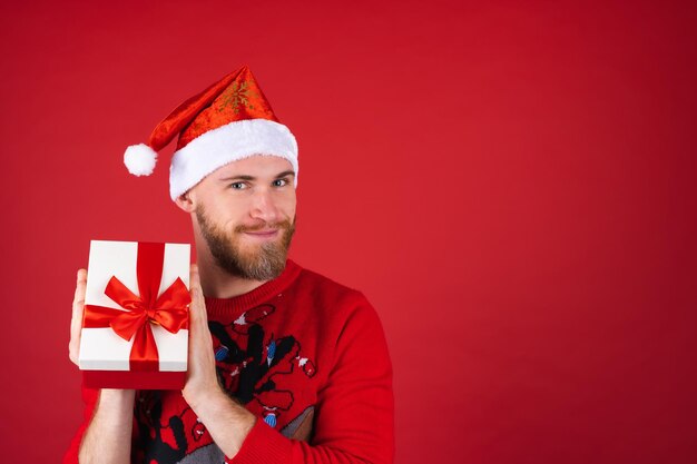 L'homme à la barbe rouge dans un chapeau de père Noël et un pull de Noël avec une boîte-cadeau est excité, impatient de passer les vacances