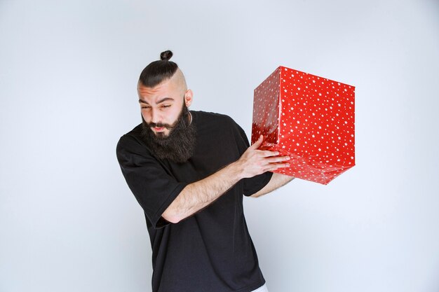 Homme à la barbe tenant une boîte-cadeau rouge et a l'air confus et terrifié.