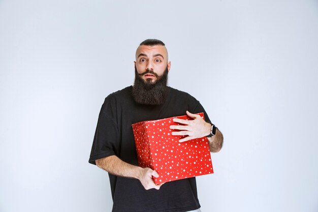 Homme à la barbe tenant une boîte-cadeau rouge et a l'air confus et terrifié.