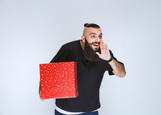 Homme à la barbe tenant une boîte-cadeau rouge et appelant quelqu'un ou chuchotant.