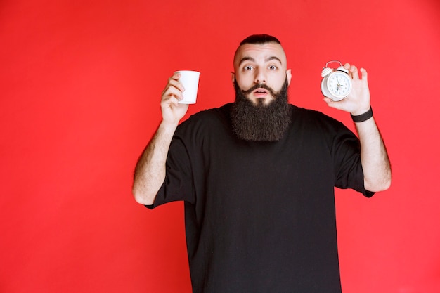 Homme à la barbe tenant un réveil et une tasse de café.