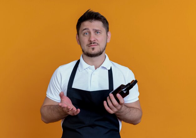 Homme barbier en tablier holding spray à la camerta à avec le bras avec une expression sceptique debout sur un mur orange