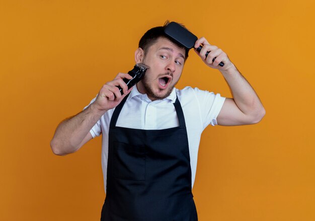 Homme de barbier en tablier raser sa barbe avec tondeuse peigner ses cheveux debout sur un mur orange