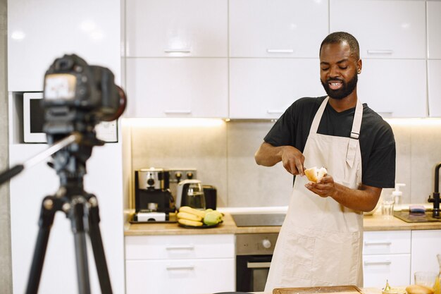 Homme barbu afro-américain souriant et cuisinant. Vidéo de tournage d'un blogueur pour cuisiner un vlog dans la cuisine à la maison. Homme portant un tablier.
