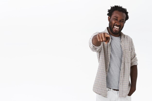 Homme barbu afro-américain sympathique et insouciant en chemise, t-shirt, pointant vers l'avant et souriant