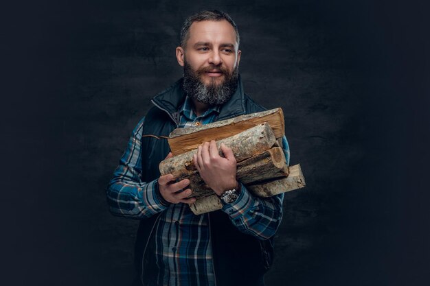 Un homme barbu d'âge moyen vêtu d'une chemise en flanelle à carreaux tient des bois de chauffage sur un fond sombre.