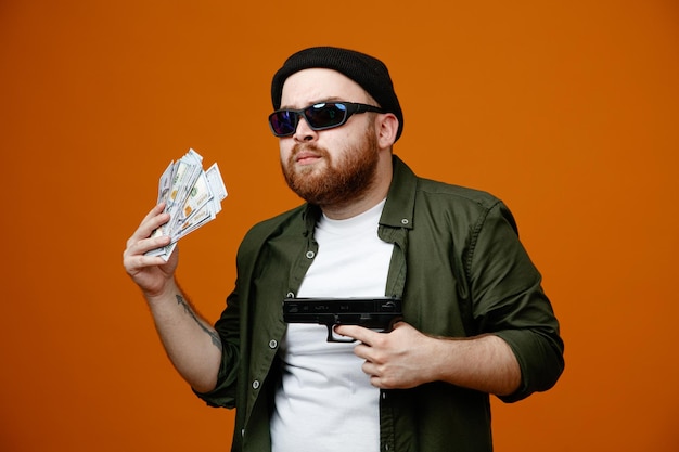 Homme barbu à l'air dangereux portant des lunettes noires et un chapeau tenant un pistolet et de l'argent à la confiance debout sur fond orange