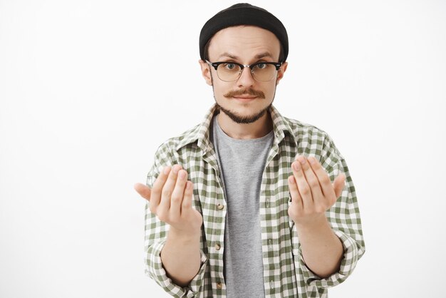 Homme barbu artistique habile à la recherche sérieuse dans des lunettes et bonnet noir levant les paumes près du corps lui demandant de s'approcher ou de lui donner quelque chose qui veut tenir l'article dans les mains sur un mur gris