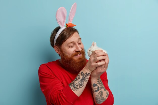 Un homme barbu au gingembre bienveillant joue avec un petit lapin mignon, porte des oreilles de lapin et un pull rouge, célèbre Pâques, apprécie le printemps, pose à l'intérieur. Concept de traditions et de fêtes religieuses