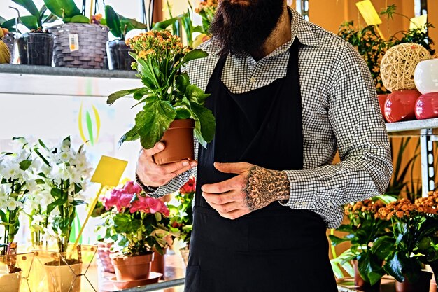 Un homme barbu aux bras tatoués tient un pot de fleurs.