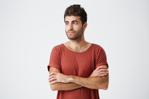 Homme barbu bronzé attrayant ayant l'air heureux debout les mains croisées contre le mur blanc. Étudiant portant une chemise t-shirt décontractée en attente de sa petite amie